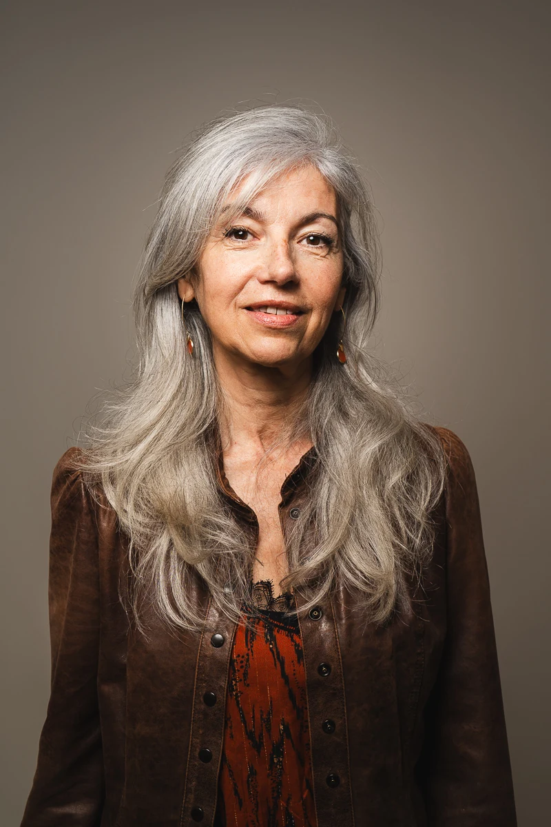 Portrait couleur d'une cheffe d'entreprise aux longs cheveux gris argentés, veste en cuir marron, léger sourire sur fond taupe