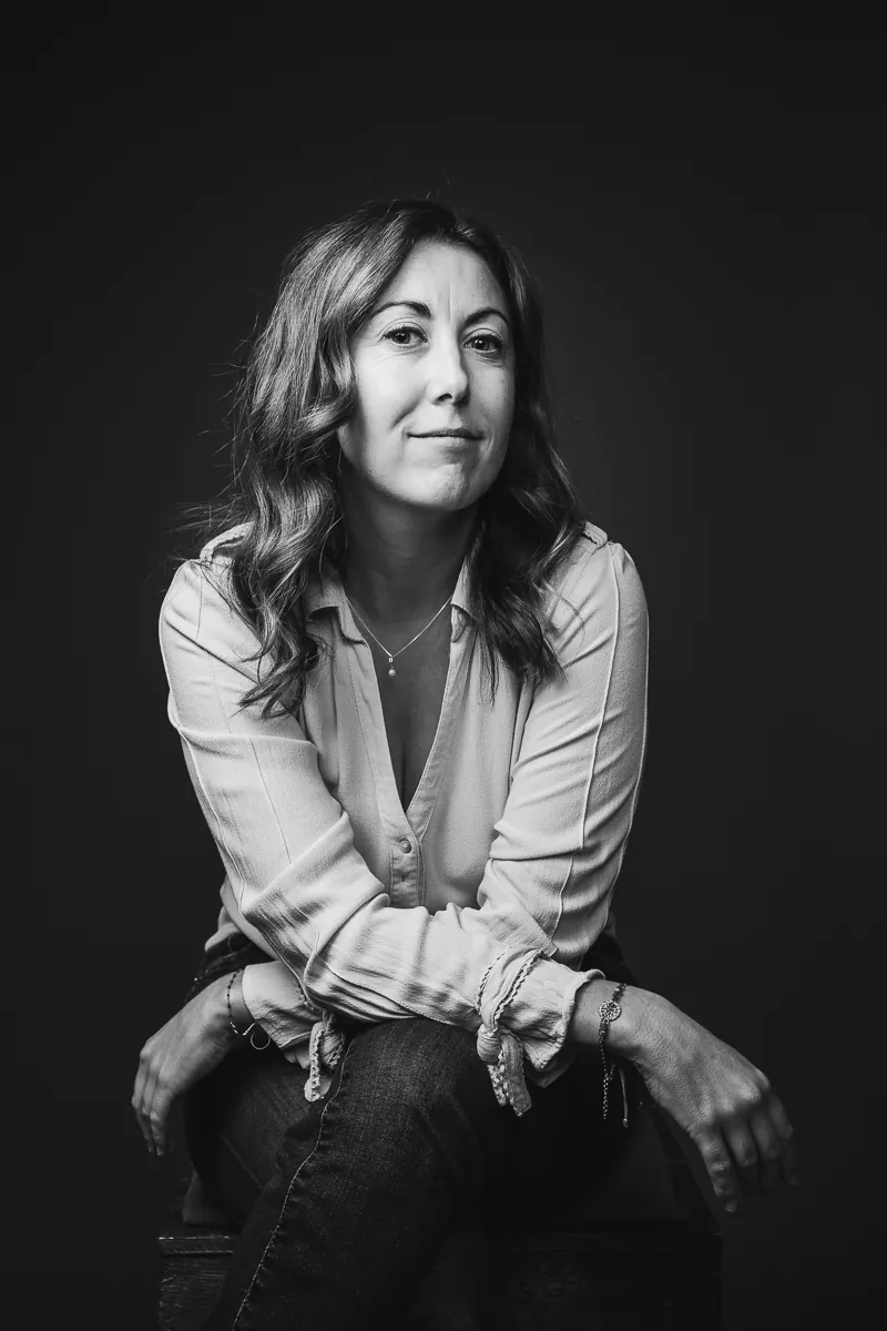 Portrait noir et blanc de Sabrina Patalano, DRH externalisée, assise, bras croisés sur les genoux, léger sourire, sur fond sombre