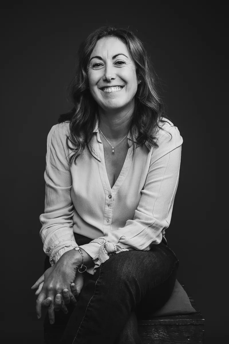 Sabrina Patalano éclate de rire, mains jointes sur les genoux, portrait noir et blanc en studio