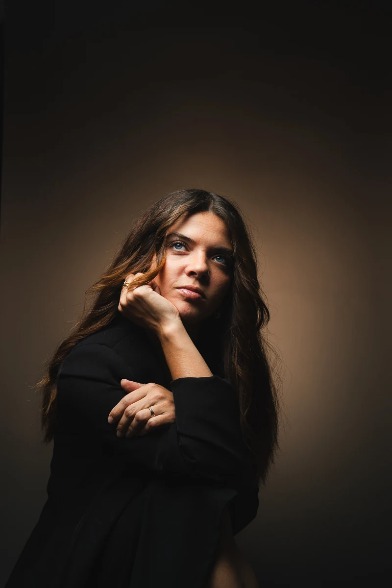 Portrait couleur d'une jeune femme aux longs cheveux bruns, la main près du visage, regard intense vers l'objectif, fond brun chaud, pour la création de contenu