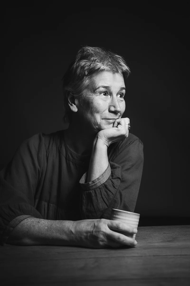 Portrait noir et blanc d'une femme accoudée à une table en bois, une tasse à la main, souriante, le regard tourné sur le côté