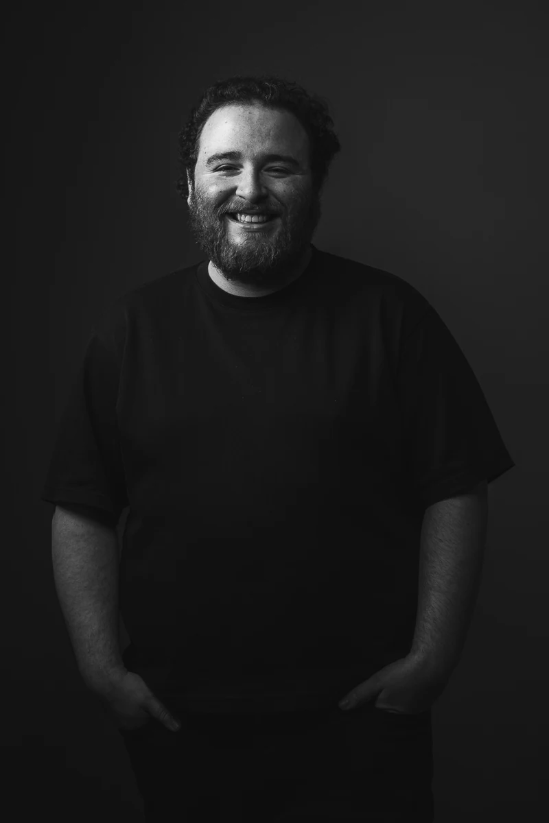 Portrait noir et blanc d'un réalisateur, debout en t-shirt noir, les mains dans les poches, grand sourire sur fond sombre