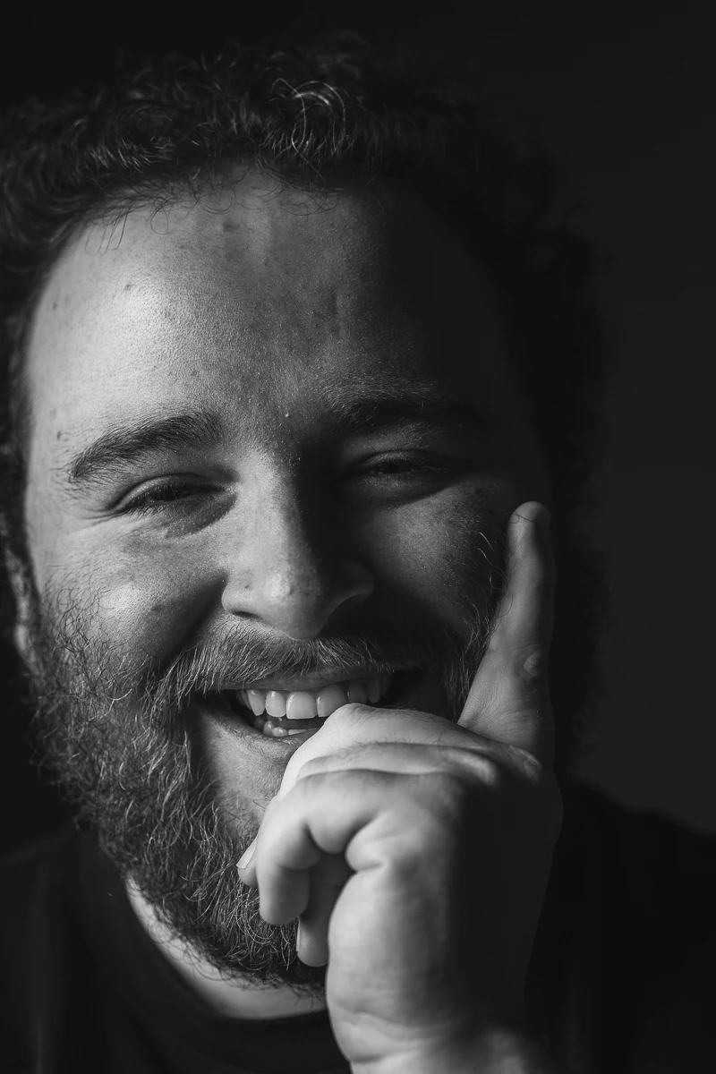 Gros plan noir et blanc d'un réalisateur en éclat de rire, la main contre la joue, sur fond noir